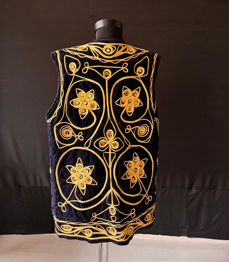 May include: A navy blue vest with intricate gold embroidery. The vest features a floral and geometric design, with circular and star-shaped elements. The gold embroidery stands out against the dark blue fabric, creating a striking contrast. The vest is displayed on a mannequin.