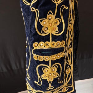 May include: Navy blue velvet vest with elaborate gold embroidery. The vest features floral and geometric designs, with small mirrored accents. The vest has a pocket and is displayed on a mannequin. The vest is a unique piece of clothing.