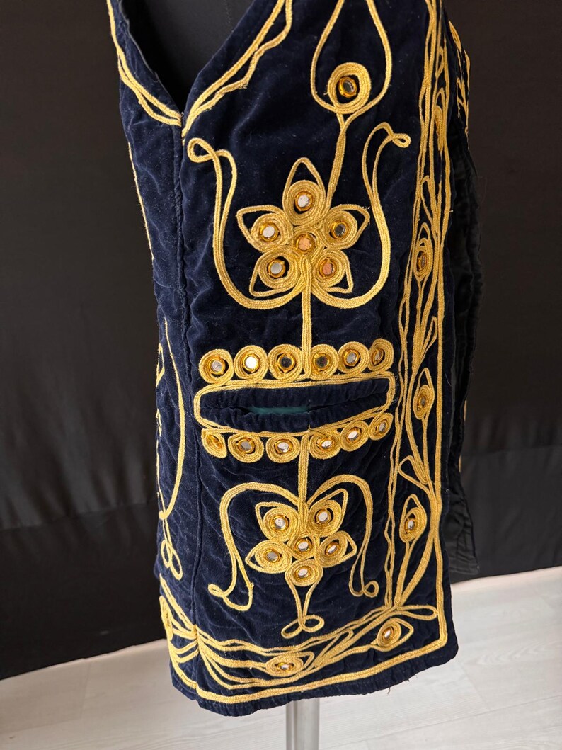 May include: Navy blue velvet vest with elaborate gold embroidery. The vest features floral and geometric designs, with small mirrored accents. The vest has a pocket and is displayed on a mannequin. The vest is a unique piece of clothing.