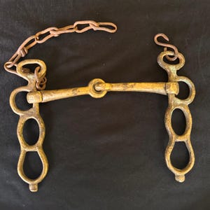 May include: An antique horse bit made of brass with a rusty chain. The bit has a straight bar with a ring in the center and decorative, elongated oval shapes on each side. The metal has a tarnished, aged appearance.