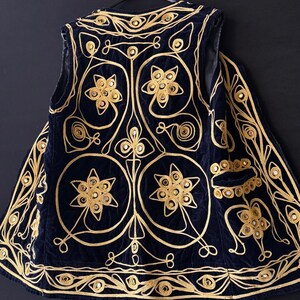 May include: A navy blue vest with intricate gold embroidery. The vest features floral and geometric patterns, with small mirrored accents. The design is symmetrical, with a detailed border and a pocket on the side. The vest is hanging on a black hanger.