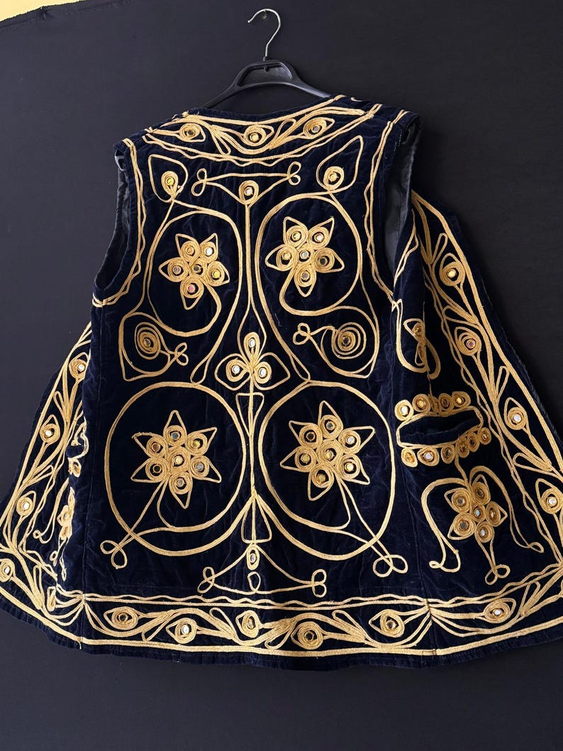 May include: A navy blue vest with intricate gold embroidery. The vest features floral and geometric patterns, with small mirrored accents. The design is symmetrical, with a detailed border and a pocket on the side. The vest is hanging on a black hanger.