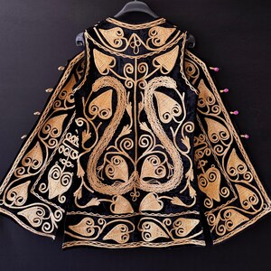 May include: Black velvet jacket with gold embroidery. The jacket has detailed floral and leaf patterns, a high collar, and long sleeves. The gold embroidery provides a striking contrast against the dark background.