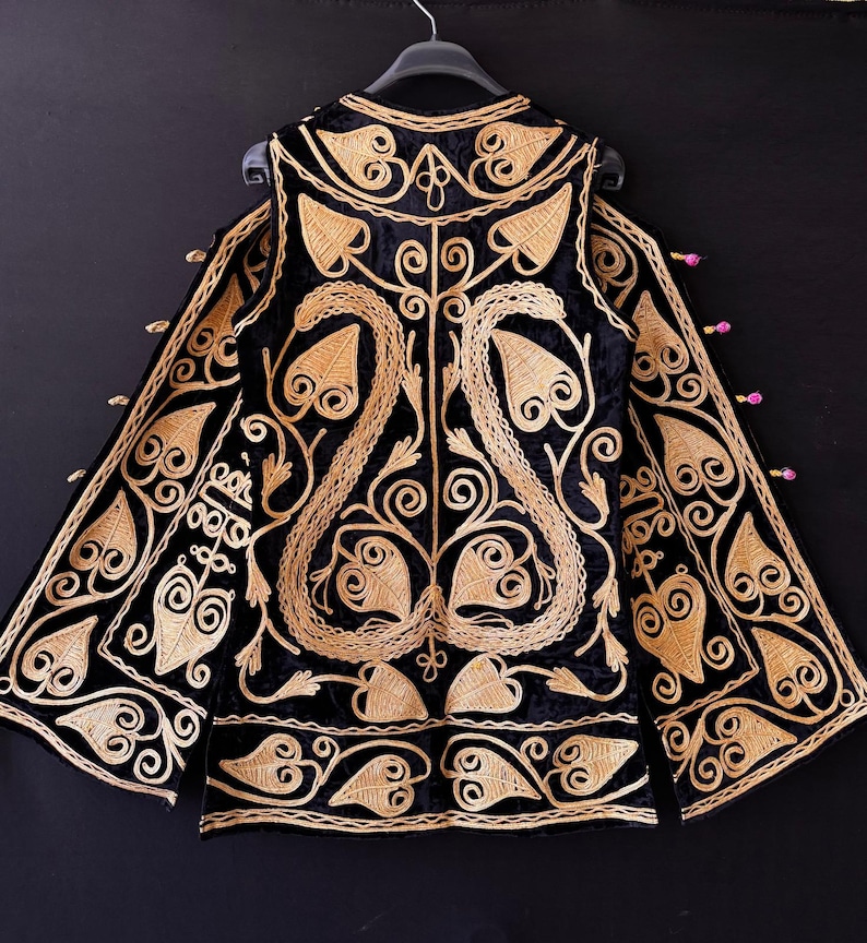 May include: Black velvet jacket with gold embroidery. The jacket has detailed floral and leaf patterns, a high collar, and long sleeves. The gold embroidery provides a striking contrast against the dark background.