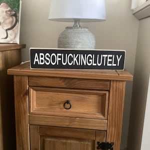 May include: A black wooden sign with white text that reads "ABSOFUCKINGLUTELY". The sign is sitting on a wooden table.