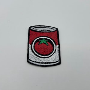 May include: Embroidered patch featuring a red tomato soup can design. The can has a white and black border, with a red tomato and green stem detail in the center. A fun, food-themed patch.