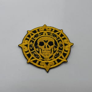 May include: Embroidered patch featuring a gold skull and a circular design with a black background. The skull has detailed eye sockets and teeth. The patch has a pointed edge and a star-like design around the circle.