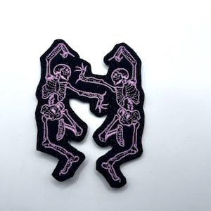 Puede incluir: Dos parches negros bordados con un diseño de esqueleto bailando. Los esqueletos están delineados en rosa y están en una pose juguetona. Los parches están sobre un fondo blanco.