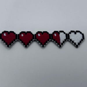 May include: A pixelated patch featuring a health bar design with five connected hearts. The hearts are red and white, with a black outline. The design is reminiscent of classic video games.
