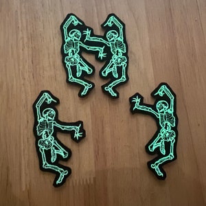 May include: Three black patches with a glow-in-the-dark skeleton design. Two patches feature two skeletons dancing, while the third patch shows a single skeleton in a dancing pose. The patches are on a wooden surface.