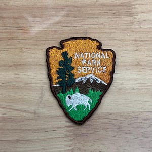 May include: A brown, green, and white embroidered patch with the National Park Service logo. The logo is a green arrowhead with a mountain range, a pine tree, and a white buffalo inside. The words "National Park Service" are written in white on a yellow background.