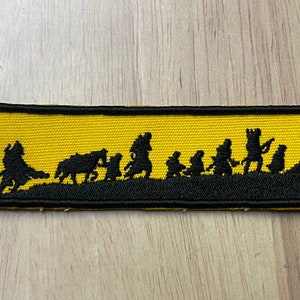 May include: A yellow embroidered patch with a black silhouette of a group of people walking, including a person riding a horse, and a person holding a staff.