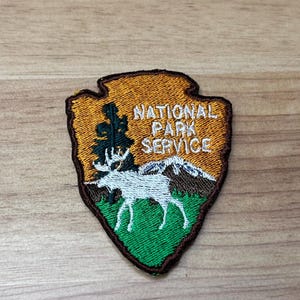 May include: Embroidered patch featuring the National Park Service arrowhead logo. The patch has a brown border and a gold background with the text "NATIONAL PARK SERVICE." The design includes a tree, mountains, and a white moose on a green field.