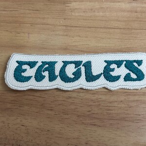 May include: White fabric patch with the word "Eagles" embroidered in teal blue lettering.