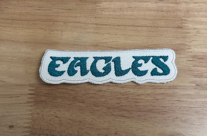 May include: White fabric patch with the word "Eagles" embroidered in teal blue lettering.