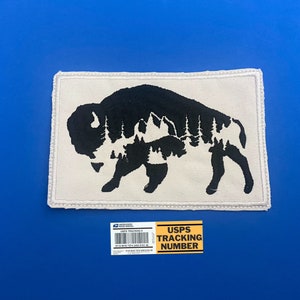 May include: A white patch with a black silhouette of a bison with a forest inside. The patch has a stitched border.