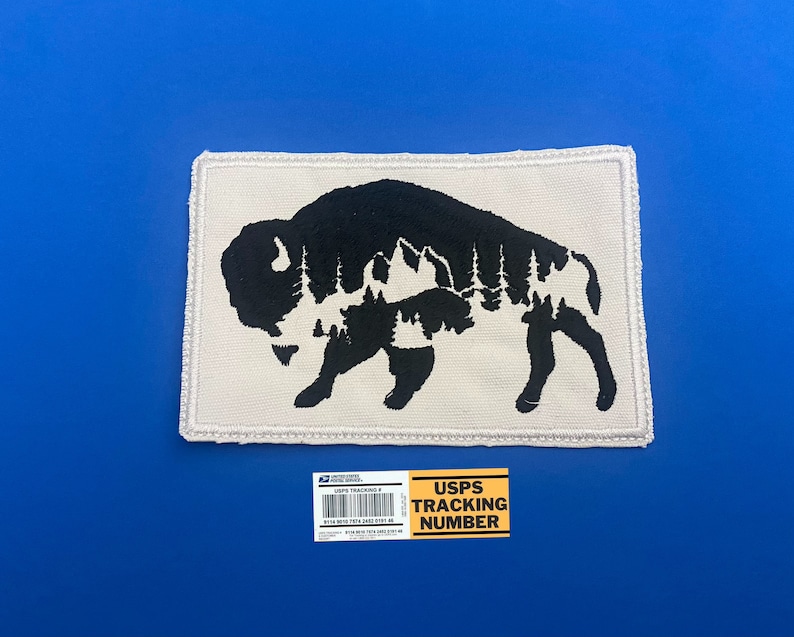 May include: A white patch with a black silhouette of a bison with a forest inside. The patch has a stitched border.