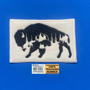 May include: White felt patch with a black embroidered silhouette of a bison with a forest inside. The patch has a white border.