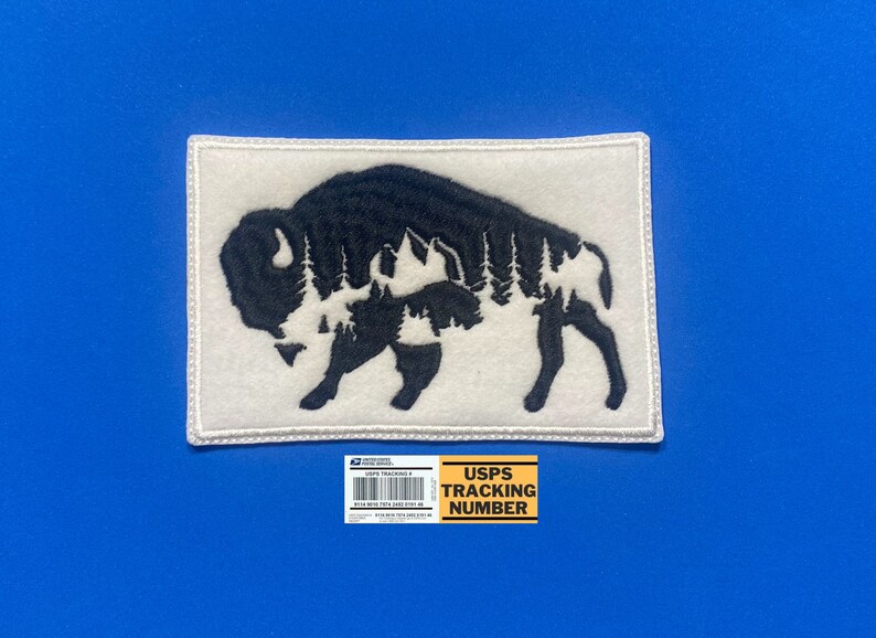 May include: White felt patch with a black embroidered silhouette of a bison with a forest inside. The patch has a white border.