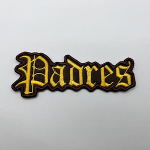 May include: Embroidered patch featuring the word "Padres" in a gothic font. The text is in gold thread, outlined in brown, and set against a brown background. The patch has a decorative border and is likely for apparel.