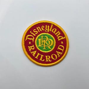 May include: Embroidered patch with the words "Disneyland Railroad" in a circular design. The patch features a red background with yellow and green accents. The center has a green circle with the letters "DR" in yellow.