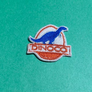 May include: Embroidered patch featuring a blue dinosaur silhouette within a white and orange circular design. The word "DINOCO" is displayed in orange block letters below the dinosaur image. The patch is on a teal background.