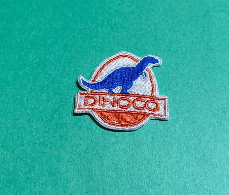 May include: Embroidered patch with a blue dinosaur silhouette inside a white and orange circular design. The word "DINOCO" is in orange block letters below the dinosaur image. The patch is set against a teal background.