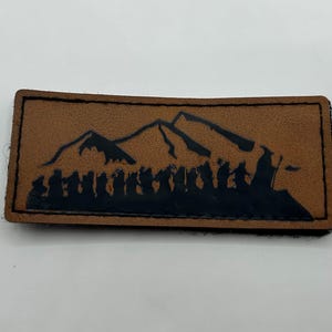 May include: A rectangular tan patch with a dark brown stitched border. The center features a black silhouette graphic of a line of figures walking in front of a mountain range. The design depicts a group of people traveling together in a landscape.