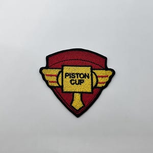 May include: Embroidered patch featuring the words "PISTON CUP" in yellow on a gold cup design with wings, set against a red shield shape. The patch has a black border and is likely for a jacket or bag.
