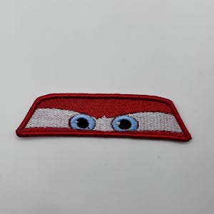 May include: Embroidered patch featuring a stylized design of car eyes. The patch has a red border, white area, and two blue eyes with black pupils. The patch is rectangular with angled sides.