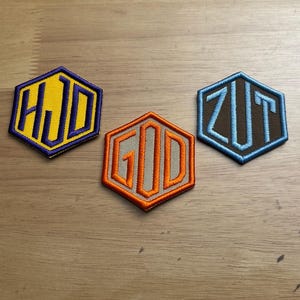 May include: Three embroidered patches in a hexagonal shape. One patch has the letters "HJD" in yellow on a purple background. Another has "GOD" in orange on a gray background. The third patch has "ZUT" in light blue on a brown background.