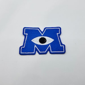 May include: A blue embroidered patch in the shape of the letter "M" with a white eye and black pupil in the center. The patch has a white border and is set against a white background.
