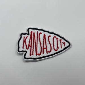 May include: Embroidered patch in the shape of an arrowhead, featuring the text "KANSAS CITY" in red stitching on a white background. The patch has a black border and is likely for apparel or accessories.