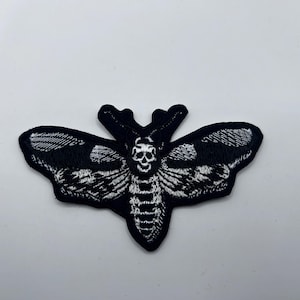 May include: Black and white embroidered patch featuring a Death's-head Hawkmoth design. The moth has a skull-like marking on its thorax and detailed wing patterns. This patch is suitable for jackets, bags, or other fabric items.