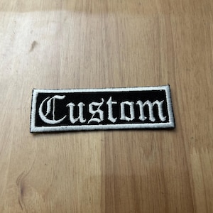 May include: Black and white embroidered patch with the word "Custom" in white lettering.