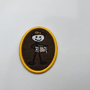 May include: Embroidered patch featuring a stick figure in a UPS uniform holding a package. The patch is oval-shaped with a brown background and a gold border. The UPS logo is on the figure's hat.