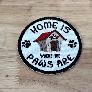 May include: White embroidered patch with black trim, featuring a cartoon dog house with a red roof and the text "Home is where the paws are".