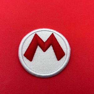 May include: A circular embroidered patch featuring a white background with a thick white border and a bold red letter M centered on the surface. The stitching is dense and uniform, creating a textured appearance for the letter and the circular base.
