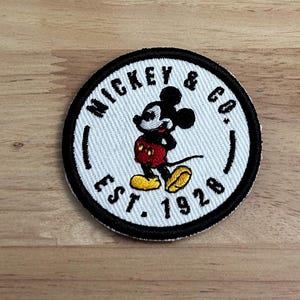 May include: White embroidered patch with a black outline featuring Mickey Mouse in a red and yellow outfit. The text "Mickey & Co. Est. 1928" is written in a black circular border around Mickey Mouse.