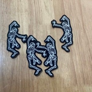 Puede incluir: Cuatro parches negros con dise&ntilde;os de esqueletos blancos en pose de baile. Los parches est&aacute;n sobre una superficie de madera marr&oacute;n claro.