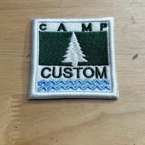 May include: White embroidered patch with a green background and a white pine tree. The text "CAMP CUSTOM" is embroidered in white on the patch. The bottom of the patch has a blue wavy line.