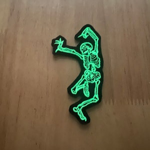 Puede incluir: Un parche negro con un esqueleto verde brillante en pose de baile. Los huesos del esqueleto est&aacute;n detallados con un color verde ne&oacute;n brillante, y el parche tiene un borde negro. Este parche es adecuado para temas de Halloween o g&oacute;ticos.