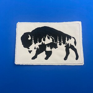 May include: A white fabric patch with a black silhouette of a bison. The bison is outlined with a white stitch. The silhouette of the bison is filled with a black and white pattern of trees and mountains.