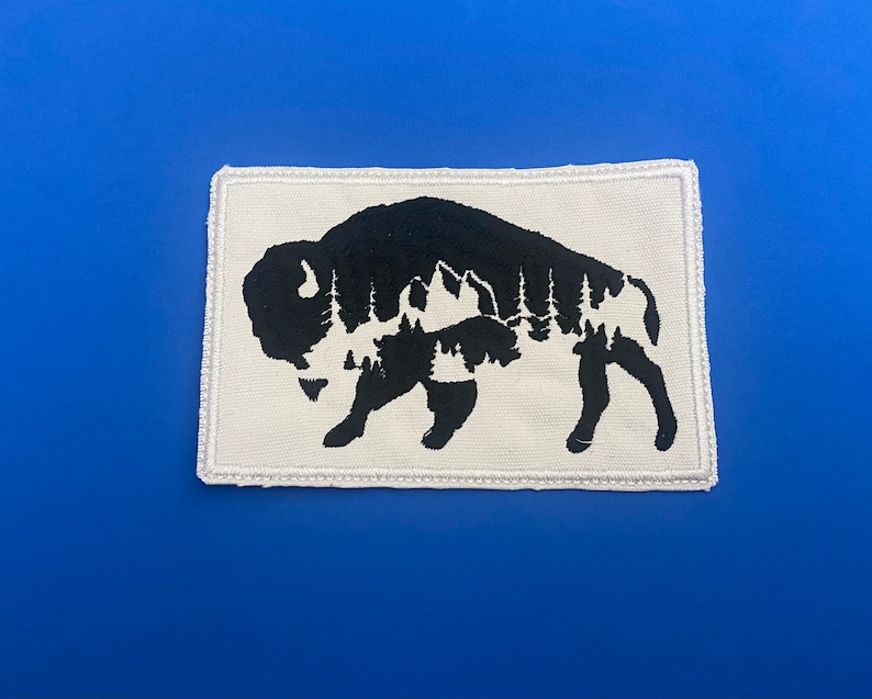 May include: A white fabric patch with a black silhouette of a bison. The bison is outlined with a white stitch. The silhouette of the bison is filled with a black and white pattern of trees and mountains.