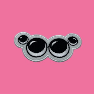 May include: Embroidered patch featuring a cartoon-style pair of eyes. The eyes are black with white highlights and a white outline, set against a light gray background. The patch is on a pink background.