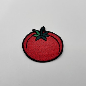 May include: Embroidered patch featuring a red tomato with green leaves and a black outline. The patch is a vibrant red color and has a detailed design. This patch is suitable for clothing, bags, or other fabric items.