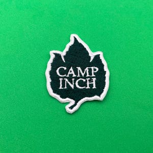 May include: A dark green embroidered patch in the shape of a leaf with the words "CAMP INCH" in white lettering. The patch has a white border and is set against a solid green background.