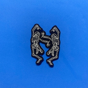 May include: Two black embroidered skeleton figures dancing against a blue background.