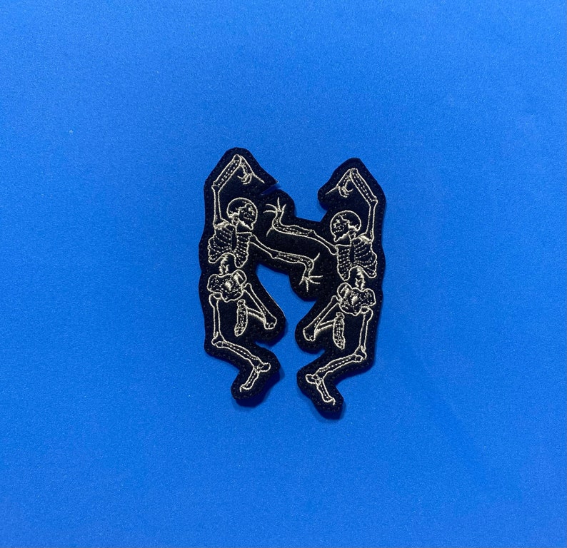May include: Two black embroidered skeleton figures dancing against a blue background.