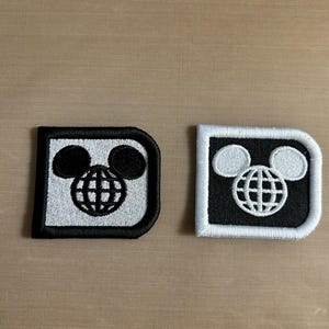 May include: Two square embroidered patches with rounded right corners. One patch features a light gray background with a black globe and mouse ear icon, bordered in black. The other patch has a black background with a white globe and mouse ear icon, bordered in white.
